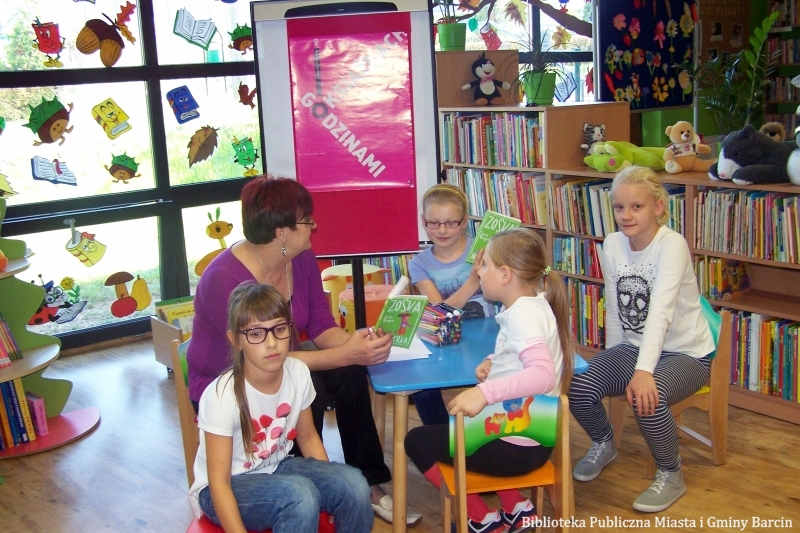4 dzieci i bibliotekarka siedzą przy stoliku, bibliotekarka trzyma w rękach ksiażki