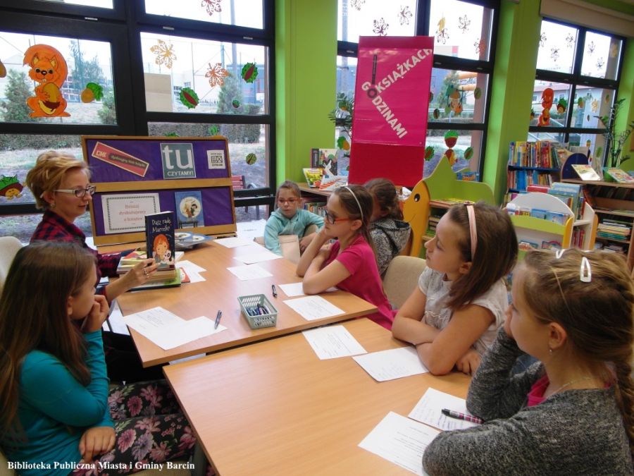6 dzieci razem z pracownicą biblioteki prowadzącą spotkanie siedzą przy stole, prowadząca pokazuje dzieciom ksiażkę.