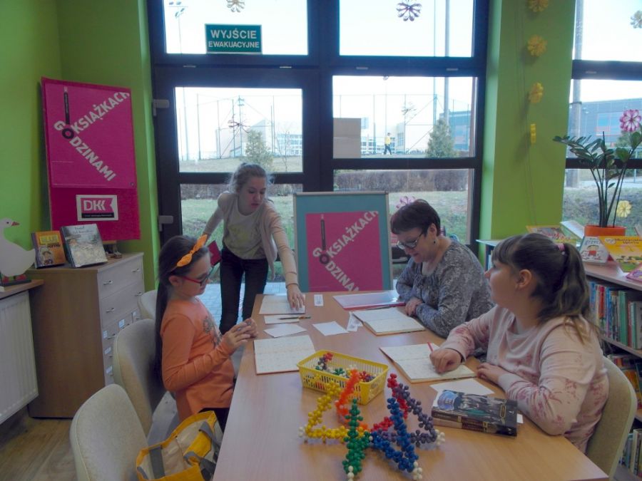 3 dziewczynki i bibliotekarka w czasie spotkania, dwie dziewczynki i bibliotekarka siedzą przy stole, jedna dziewczynka stoi i sięga ręką w stronę stołu.