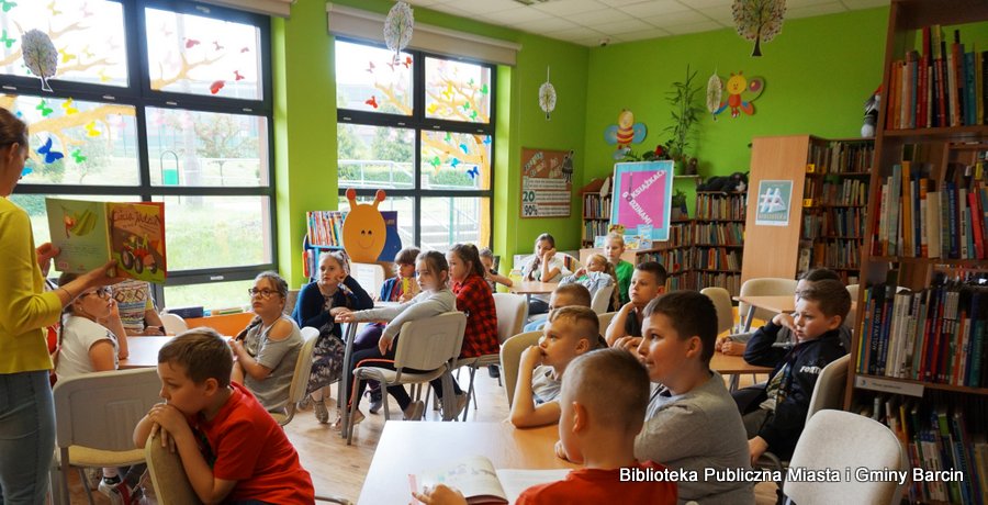 Dzieci siedzą przy stolikach, bibliotekarka prowadząca spotkanie pokazuje zebranym ksiażkę.