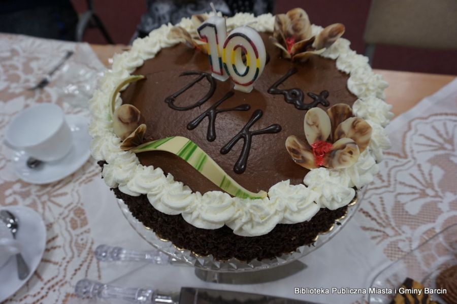 Zdjęcie tortu przygotowanego z okazji 10 rocznicy Dyskusyjnego Klubu Książki w Barcinie, jest to czekoladowy tort z ozdobiony białym kremem i kwiatkami z dwukolorowej czekolady.