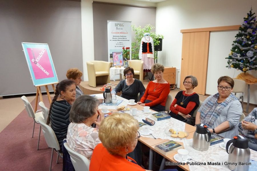 Osiem kobiet siedzi przy stołach ustawionych w rzędzie, na stołach leży poczęstunek w formie ciastek, kawy i herbaty, oraz omawiane ksiażki