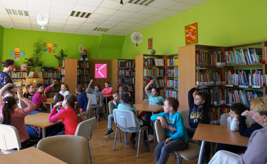 Dzieci razem z opiekunami siedzą przy stolikach pomiędzy stolikami chodzi bibliotekarka prowadząca zajęcia.