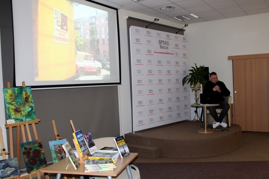 Po prawej Pan Borys Tynka siedzący w fotelu na scenie zwrócony do publiczności, po lewej wyświetlana jest prezentacja złożona ze zdjęć.