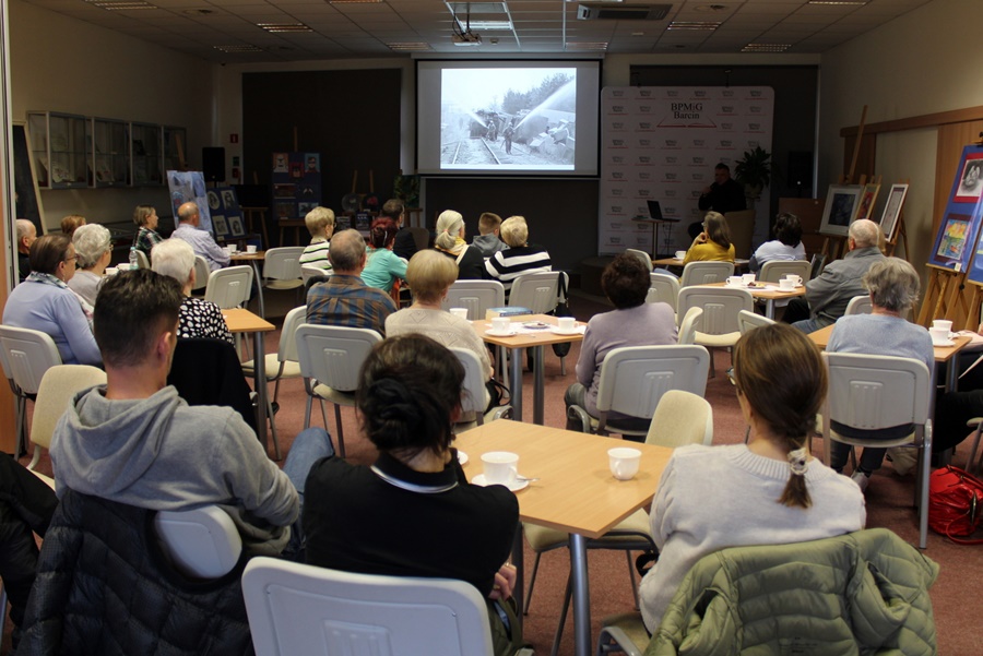 Widok zza publiczności pokazujący zebranych ludzi słuchających prowadzącego spotkanie autora i oglądających zdjęcia z prezentacji.