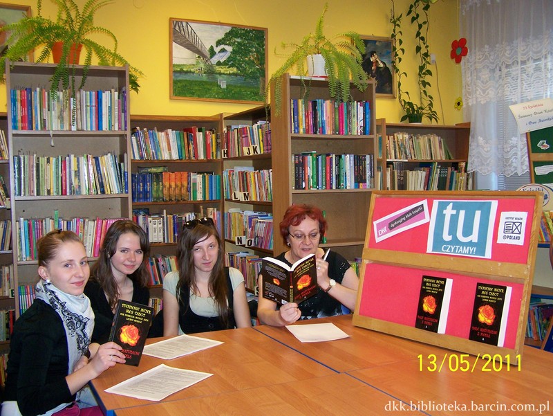 Uczestniczki spotkania razem z prowadzącą siedzą przy stołach, uczestniczki patrzą w obiektyw aparatu w tle stoją regały z ksiażkami