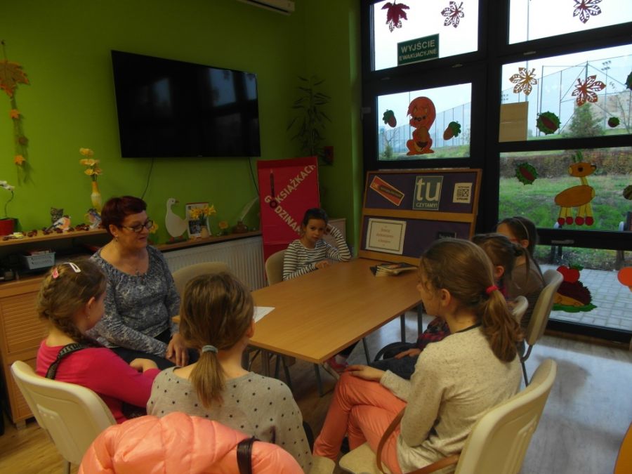 Prowadząca spotkanie bibliotekarka rozmawia z dziećmi zebranymi wokół stołu.