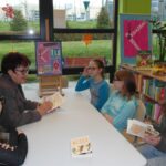 5 osób siedzących po dwóch stronach stołu, bibliotekarka i jedna z dziewczynek trzyma w rękach książkę, inne książki leżą na stole.