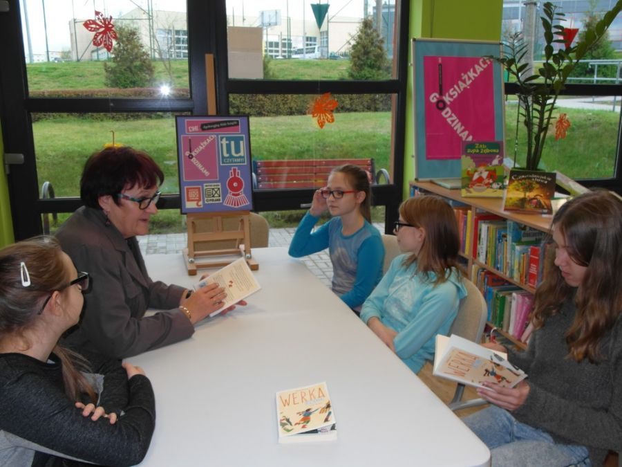 5 osób siedzących po dwóch stronach stołu, bibliotekarka i jedna z dziewczynek trzyma w rękach książkę, inne książki leżą na stole.