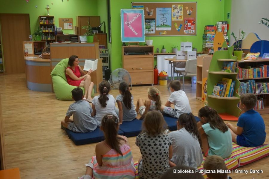 grupa dzieci siedzi na materacach i słucha czytanej przez bibliotekarkę książki.