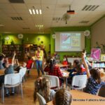 Bibliotekarka chodzi pomiędzy stolikami, dziesi siedzące przy tych stolikach podnoszą ręce zgłaszając chęć odpowiedzi na pytanie.