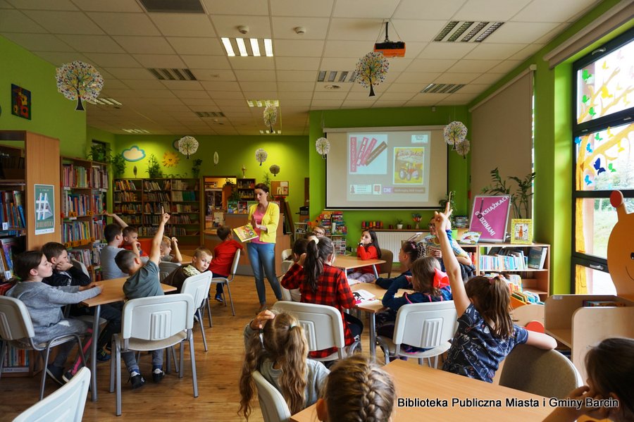 Bibliotekarka chodzi pomiędzy stolikami, dziesi siedzące przy tych stolikach podnoszą ręce zgłaszając chęć odpowiedzi na pytanie.