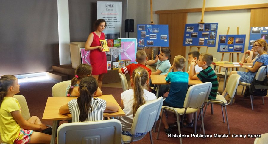 Dzieci razem z opiekunką siedzą przy stolikach, przed nimi stoi bibliotekarka trzymająca w rekach książkę.