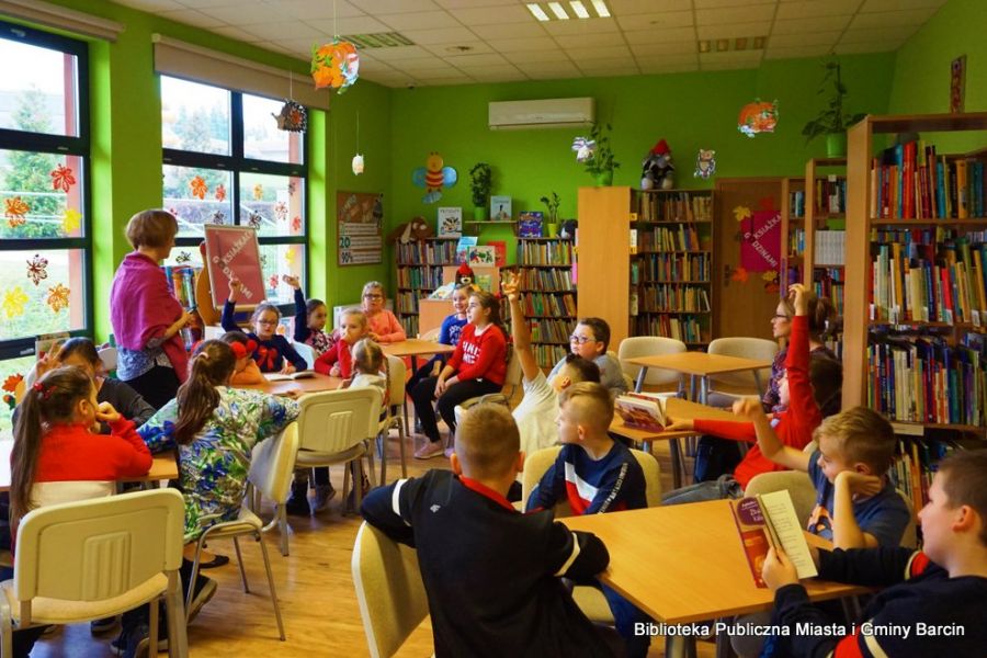 Dzieci siedzą przy stolikach, niektóre z nich podnoszą rękę do odpowiedzi, bibliotekarka chodzi pomiędzy stolikami.