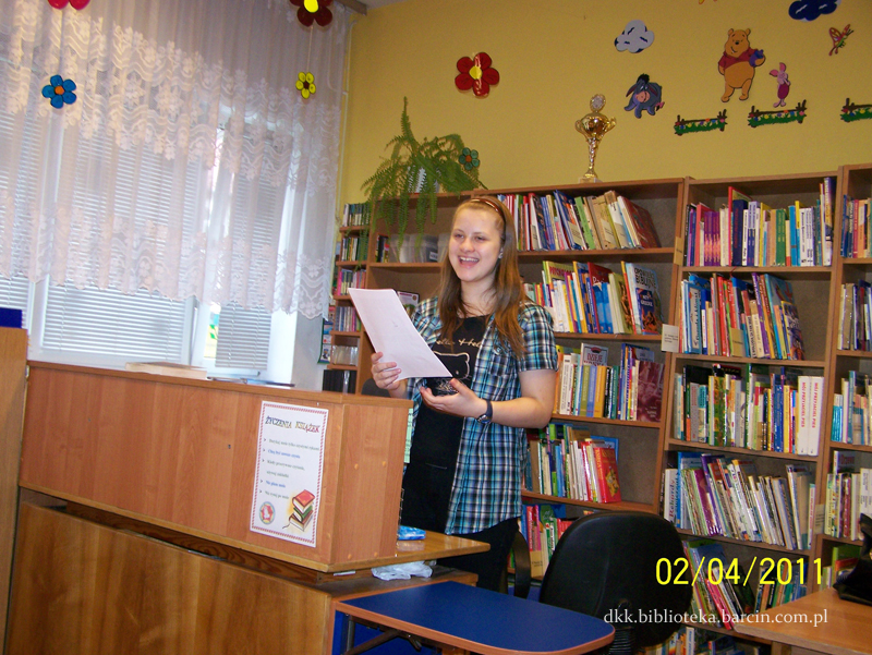 Dziewczyna o blond włosach ubrana w czarny podkoszulek, koszulę w niebieską kratę i czarne spodnie stoi za ladą biblioteczną z kartką w ręce