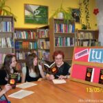 Cztery uczestniczki spotkania siedzą przy stołach, jedna z uczestniczek i prowadząca trzymają otwarte książki, z prawej strony stoi tablica z logo DKK, instytutu ksiażki i dwiema omawianymi książkami, w tle stoją regały z ksiażkami.