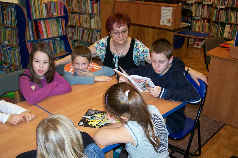 5 dzieci siedzi przy stole, jedno z dzieci czyta książkę, bibliotekarka prowadząca zajęcia przygląda się grupie.