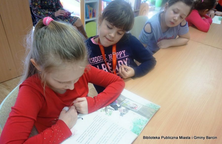Jedna z dziewczynek czyta książkę, dziewczynki siedzące obok niej patrzą w jej stronę.