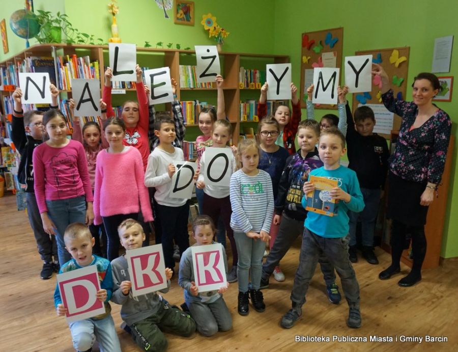 Grupowe zdjęcie uczestników spotkania, dzieci ustawione w 3 rzędach, dwa rzędy soją pierwszy rząd klęczy na podłodze, dzieci trzymają napis "Należymy do DKK", obok dzieci w trzecim rzędzie stoi bibliotekarka prowadząca spotkanie.