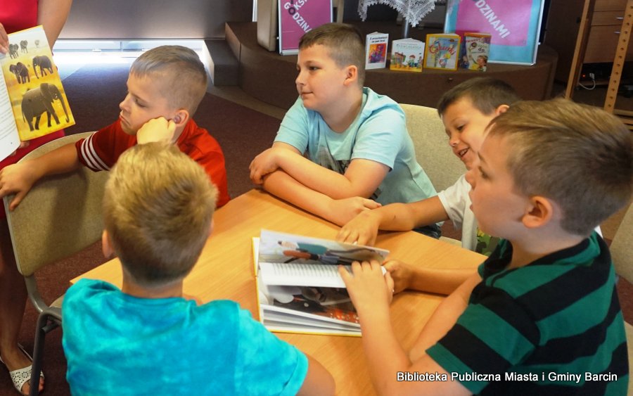 Grupa 5 chłopców siedząca przy stoliku, na stole leży otwarta książka, chłopcy jednak patrzą na książkę pokazywaną przez bibliotekarkę.