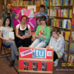 6 czytelniczek i prowadząca na grupowym zdjęciu , dwie czytelniczki trzymają plakat, z przodu stoi tablica informacyjna z logotypami i okładką książki.