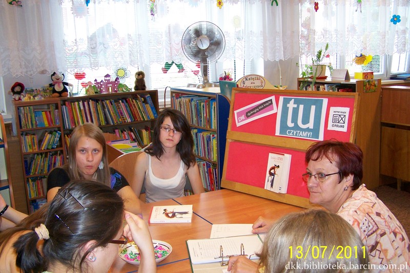 5 osób siedzi przy stołach w trakcie spotkania, prowadząca ma przed sobą otwarty segregator, na stole stoi miseczka z cukierkami i omawiana książka.