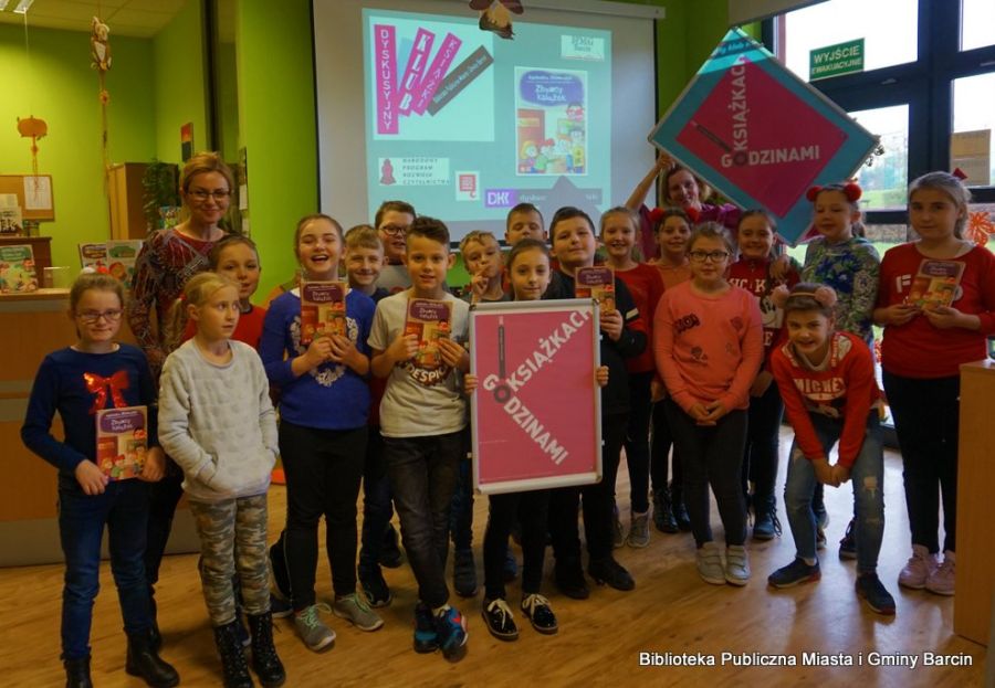 Grupowe zdjęcie uczestników spotkania, dzieci stoją w 3 rzędach, niektóre z nich trzymają książki wychowawczyni klasy i bibliotekarka stoją z tyłu za całą grupą, bibliotekarka trzyma uniesiony plakat z napisem "Godzinami o książkach".