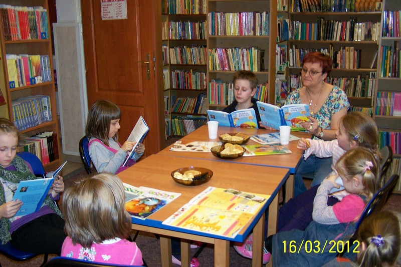 6 dzieci i bibliotekarka prowadząca spotkanie  siedzą przy stołach, 3 dzieci i bibliotekarka czytają książki trzymane w rękach, inne dzieci słuchają, jedna dziewczynka pije sok z jednorazowego kubka