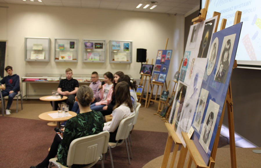 Grupa młodych osób których prace są wystawiane na wystawie, 4 dziewczyny i 2 chłopców siedzących na krzesłach, obok nich siedzi bibliotekarka prowadząca spotkanie, po prawej stronie zdjecia widać niektóre z prezentowanych prac