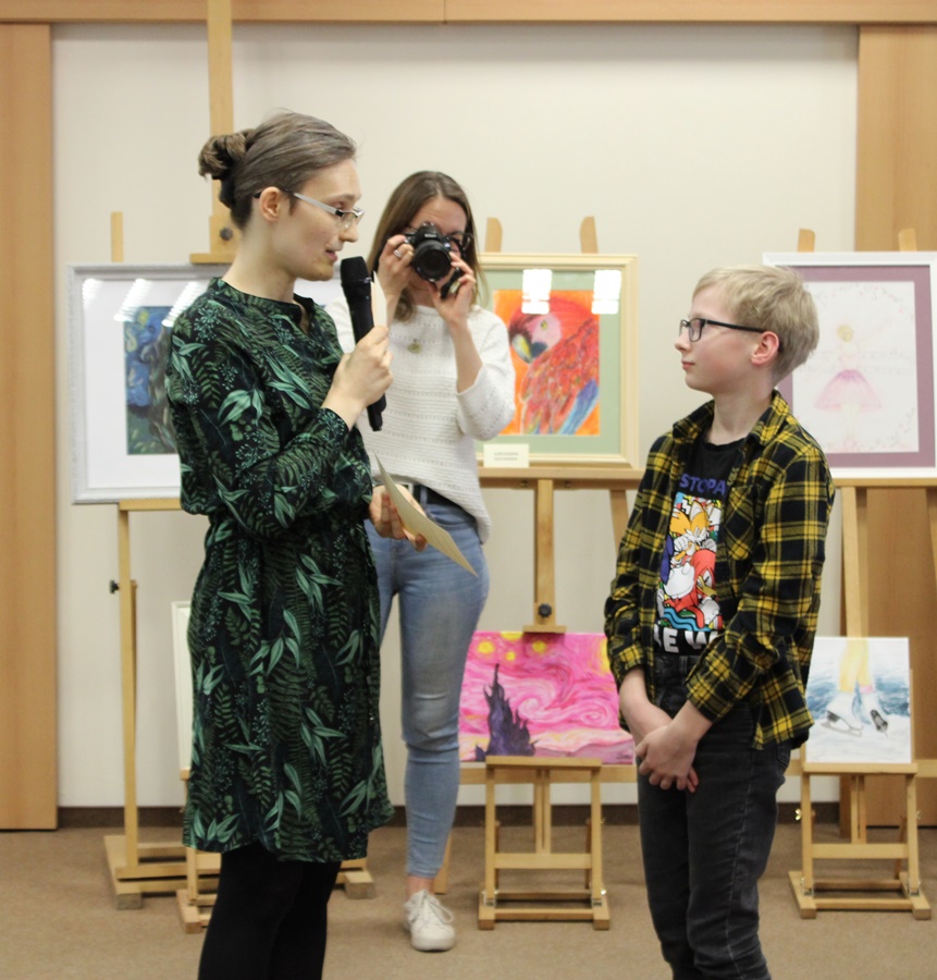 Bibliotekarka wręczająca dyplom młodemu chłopcu, którego prace są pokazywane na wystawie, w tle widać kobietę fotografującą ten moment.