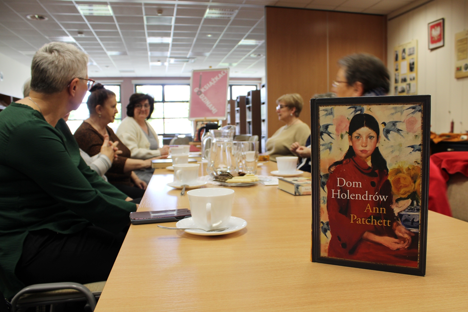 Na pierwszym planie widać książkę pt "Dom Hollendrów" Ann Patchett, w tle widać ludzi siedzących wzdłuż stołów, filiżanki do kawy, ciastka na talerzyku.