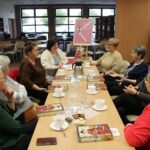 8 kobiet siedzi i rozmawia, na stołach leżą książki, stoi poczęstunek w formie kawy, herbaty i ciastek, w głębi zdjęcia widać plakat z napisem "Godzinami o ksiażkach" umiejscowionym na różowym tle.