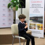 Chłopiec, uczestnik konkursu siedzący na krześle, ubrany w biemny sweter spod którego widać kołnieżyk koszuli w kolorową kratę, czarne spodnie i białe buty, w ręku trzyma mikrofon.