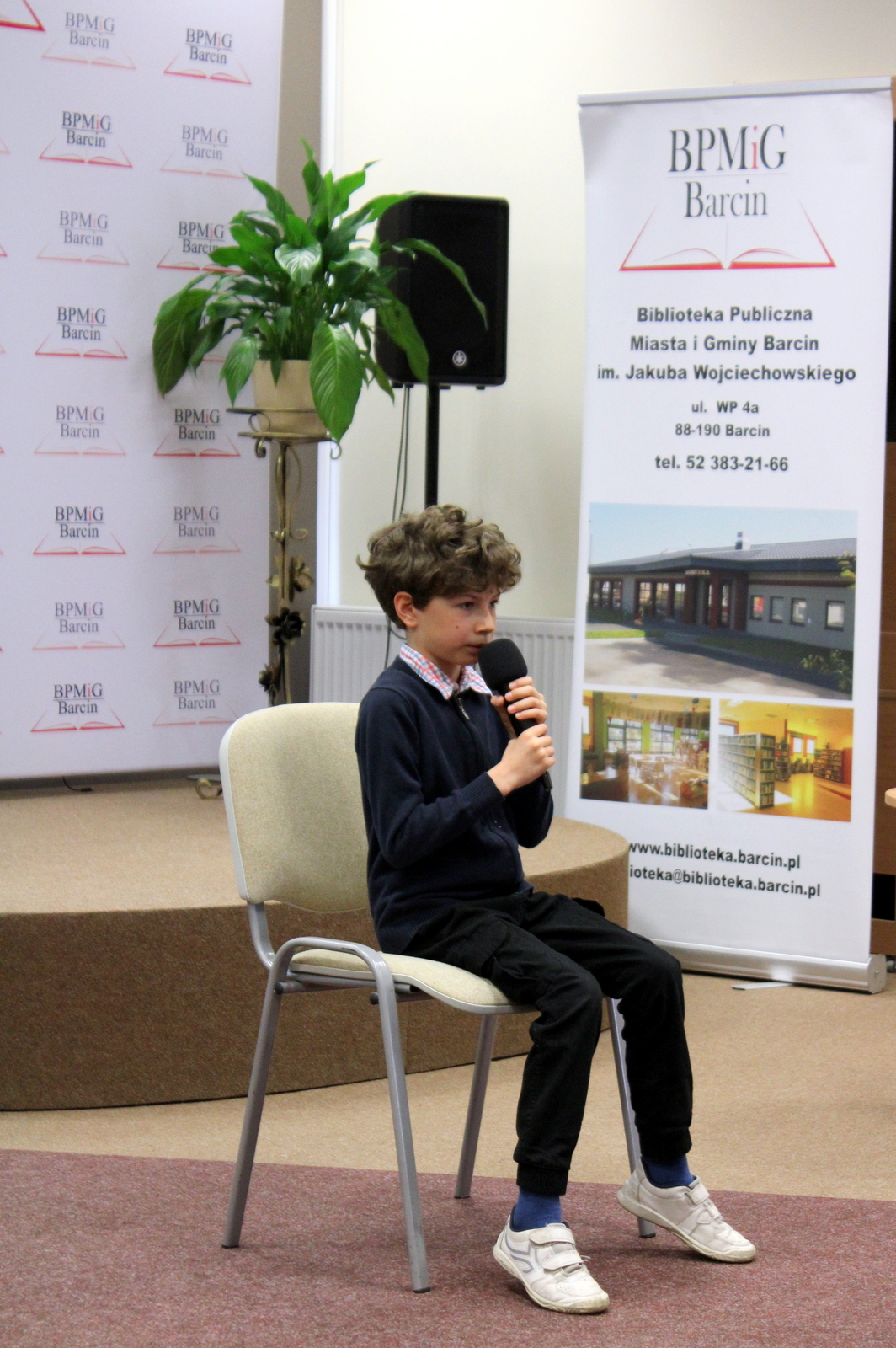 Chłopiec, uczestnik konkursu siedzący na krześle, ubrany w biemny sweter spod którego widać kołnieżyk koszuli w kolorową kratę, czarne spodnie i białe buty, w ręku trzyma mikrofon.