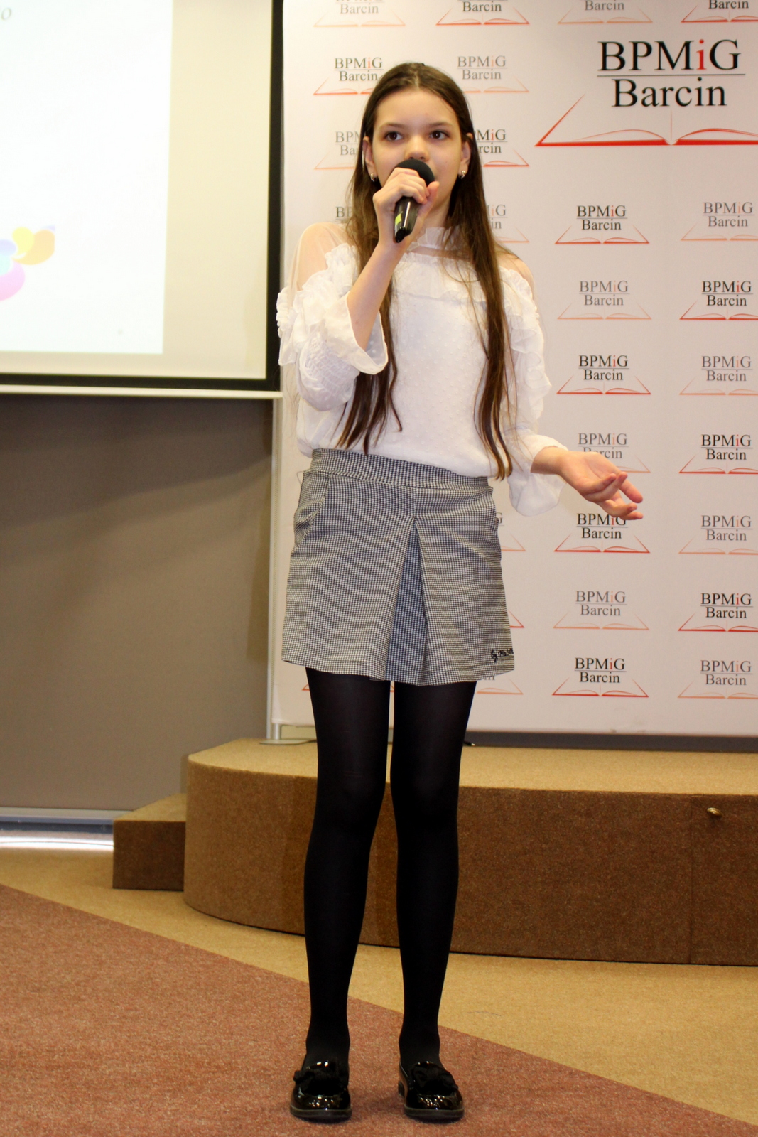 Uczestniczka konkursu, dziewczyna z ciemnymi długimi włosami ubrana w białą bluzkę, krótką spódniczkę w drobną biało czarną krate, czarne rajstopy i czarne błyszczące buty