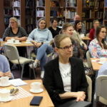 Część widowni zgromadzonej na spotkaniu, widać osoby w różnym wieku o różnej płci siedzące przy stolikach.