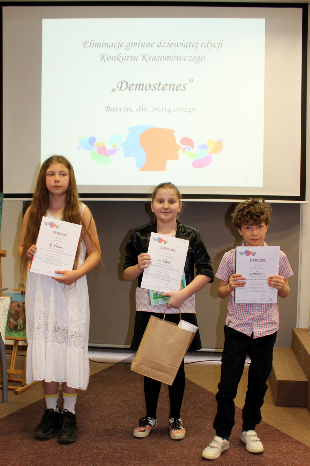 Troje uczestników konkursu trzymających wręczone nagrody oraz dyplomy, za nimi wyświetlany jest plakat informujący o konkursie.
