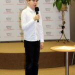 Uczestnik konkursu, chłopiec o rudych włosach ubrany w białą koszulę, czarne spodnie i czarne adidasy, trzyma w ręku mikrofon.