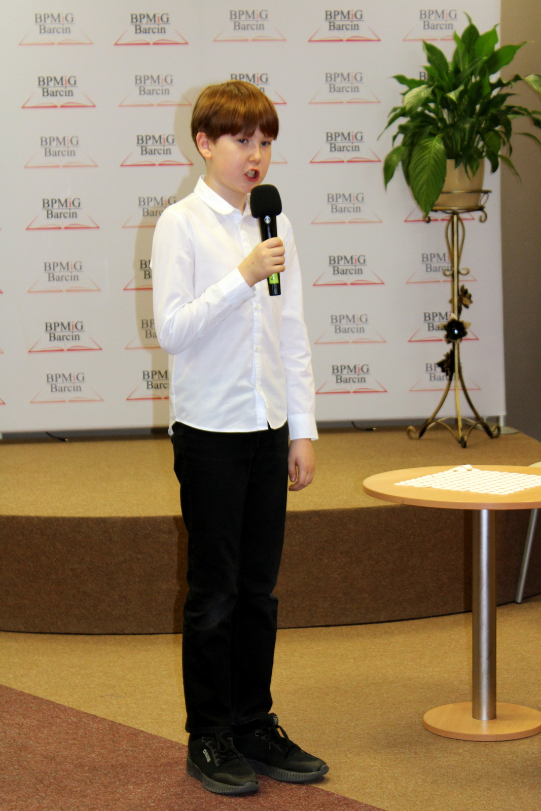 Uczestnik konkursu, chłopiec o rudych włosach ubrany w białą koszulę, czarne spodnie i czarne adidasy, trzyma w ręku mikrofon.