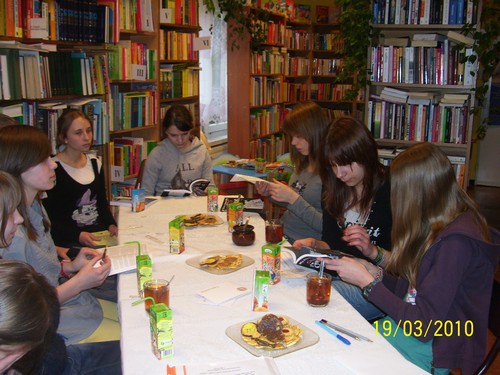 Osiem dziewczyn siedzących wokół stołu, część czyta książki, inne słuchają  lub rozmawiają