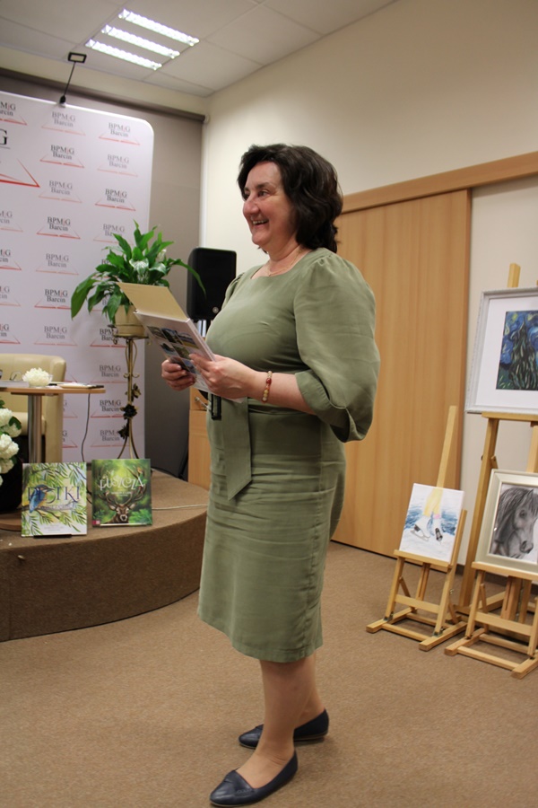 Dyrektor biblioteki, brunetka ubrana w zieloną suknię i czarne buty, na nadgarstku widać bransoletkę, stoi uśmiechnięta w rękach trzyma upominek dla zaproszonego gościa.