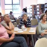 Część osób zebranych na widowni, widać ludzi siedzących pzry stolikach, w tle pracownik biblioteki nalwewa kawę jednej z osób które przyszły na spotkanie.