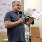 Pan Marcin Kostrzyński stoi przed publicznością i wskazuje na książkę trzymaną w jednej ręce.