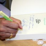 Zbliżenie na podpisywaną książkę, widać dłoń trzymającą długopis, pieczątkę i dedykację złożoną na pierwszej stronie otwartej książki.