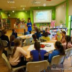 12 dzieci siedzi przy stołach, przed nimi stoi bibliotekarka, coś do nich mówi, dzieci patrzą w jeje stronę.