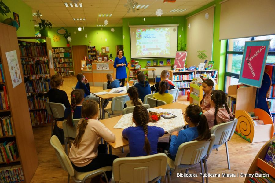 12 dzieci siedzi przy stołach, przed nimi stoi bibliotekarka, coś do nich mówi, dzieci patrzą w jeje stronę.