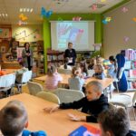 dzieci siedzą przy stolikach, nauczycielka z bibliotekarką stoją z przodu grupy.