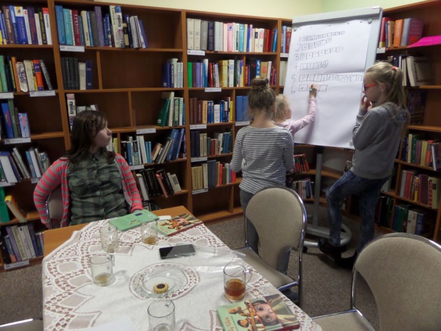 4 dziewczynki, trzy stoją przy tablicy z kartkami papieru, jedna siedzi przy stole