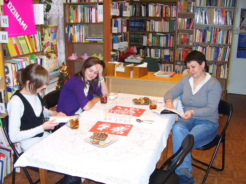 Dwie dziewczyny i bibliotekarka siedzą przy stole, dwie osoby trzymają otwarte książki