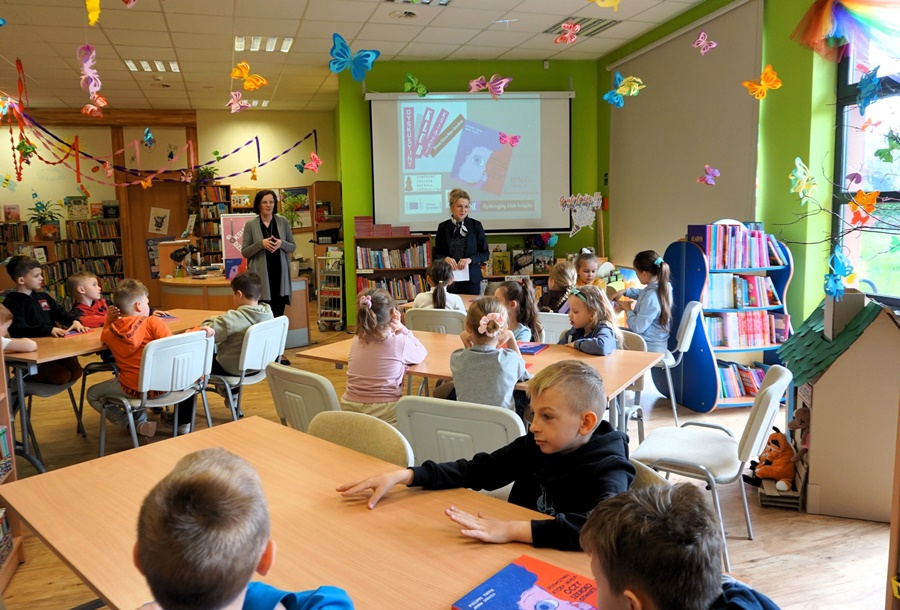 dzieci siedzą przy stolikach, nauczycielka z bibliotekarką stoją z przodu grupy.
